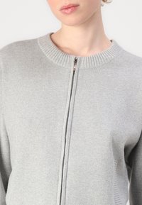 Šviesiai pilka zip-up megztinis su riloto apykakle ir tekstūra. Užtrauktukas eina vertikaliai žemyn per priekinę dalį, pabrėždamas megztos medžiagos raštą.