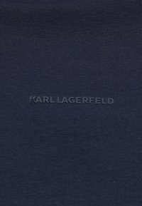 Tamnoplava tkanina s teksturiranim, rebrastim završetkom s izbočenim logotipom "KARL LAGERFELD" u kontrastnoj nijansi na sredini.