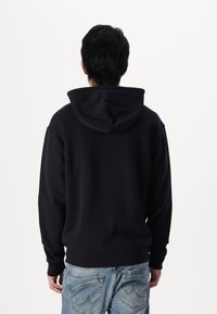 Sudadera negra con capucha hecha de tela suave, que cuenta con un bolsillo tipo canguro, puños y cintura acanalados. Se lleva con vaqueros azul claro.