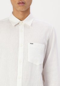Biała lniana koszula z kołnierzykiem na guziki, pojedynczą kieszenią na piersi i subtelną teksturą. Posiada logo "Wrangler" na kieszeni.