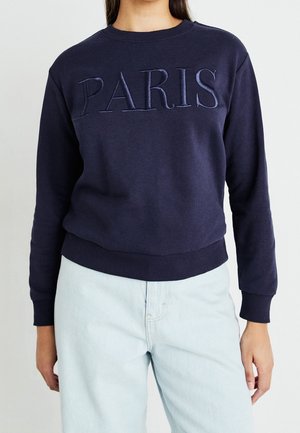 Kvinde iført mørkeblå sweatshirt med hævet "PARIS" tekst og lyseblå jeans, stående foran en ensartet lys baggrund.