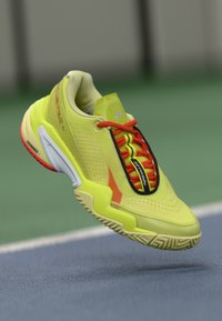 Felgele sportschoen met rode veters en zwarte details, zwevend boven het oppervlak van een tennisbaan.