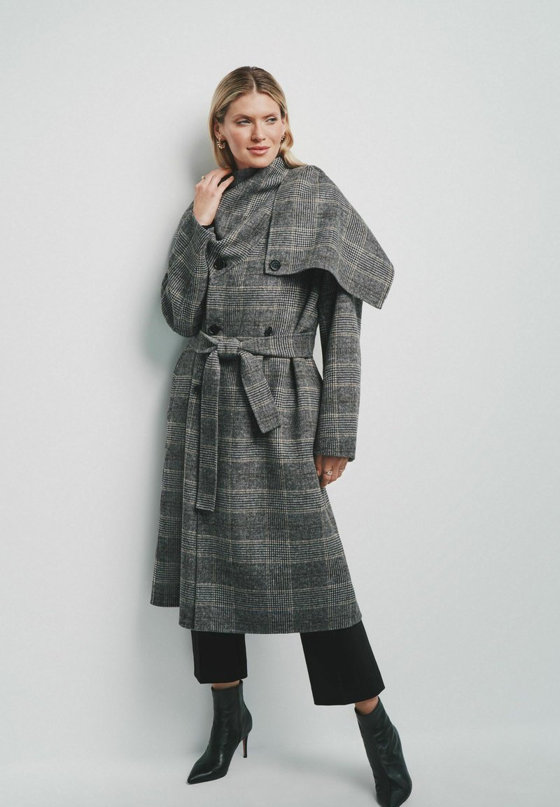 Szary płaszcz w kratę o konstrukcji zakładkowej, z podwójnymi guzikami, szerokim kołnierzem i paskiem w talii, w połączeniu z czarnymi spodami typu cropped i botkami na szpilce.