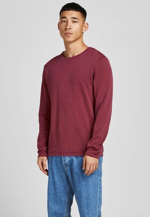 Maglietta a maniche lunghe color bordeaux con collo a giro e polsini a costine, indossata con jeans in denim blu. Il tessuto appare morbido con una texture minima.