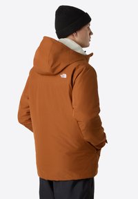 Chaqueta aislante naranja con capucha, que cuenta con un forro de felpa color crema y un pequeño logo blanco en el hombro izquierdo. Se lleva un gorro de punto negro.
