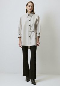 Giacca camicia beige chiaro con colletto, bottoni frontali e vita cintrata, abbinata a pantaloni neri a zampa d'elefante e stivaletti. Tessuto liscio.