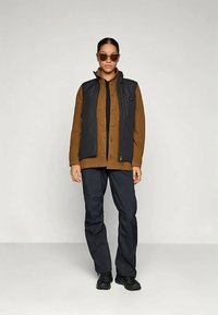 Femeie îmbrăcată cu o cămașă maro cu nasturi, un veston negru, pantaloni negri, pantofi negri și ochelari de soare maro, stând în fața unui fundal simplu.