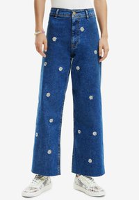 Blue denim wide-leg jeans with white daisy embroidery, high waist, and a button front. Paired with colorful patterned sneakers.