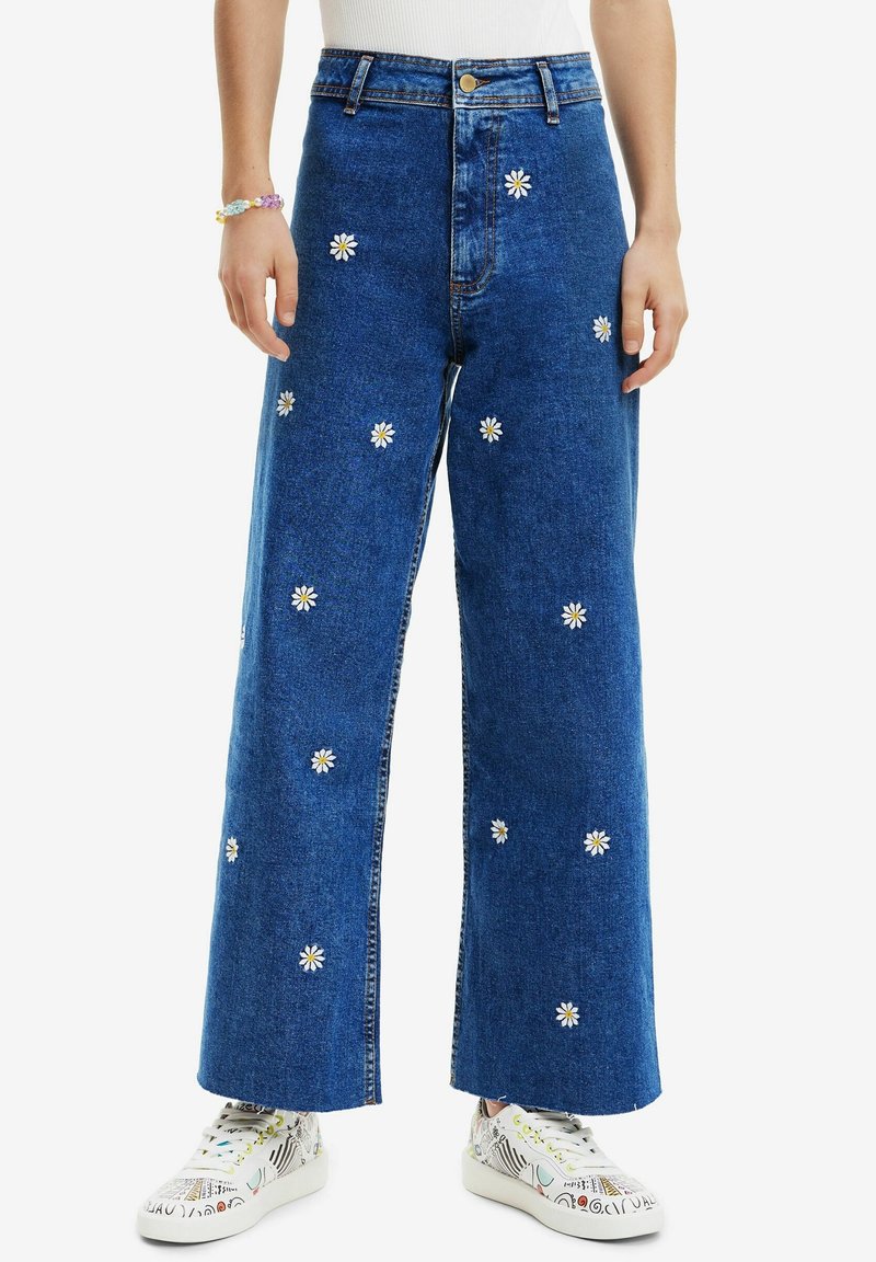 Blue denim wide-leg jeans with white daisy embroidery, high waist, and a button front. Paired with colorful patterned sneakers.