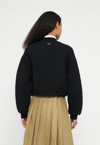 Μαύρο μπουφάν bomber με καπιτονέ ύφασμα, με ριμπ λαιμόκοψη και μανσέτες, που διαθέτει μια μικρή ετικέτα μάρκας. Φορεμένο πάνω από μια πλισέ μπεζ φούστα.