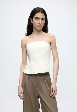 Młoda kobieta o długich ciemnych włosach, ubrana w biały top bez ramiączek z baskinką oraz brązowe plisowane spodnie z wysokim stanem na jednolitym tle.