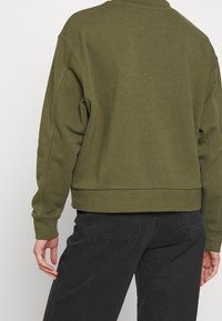 Persona vistiendo un sudadera de manga larga verde oliva y jeans negros, fotografiada desde atrás contra un fondo liso.