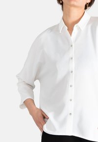 Camisa blanca de botonadura en tejido ligero, con cuello clásico, mangas largas y un ligero brillo. Los botones son de diferentes colores.