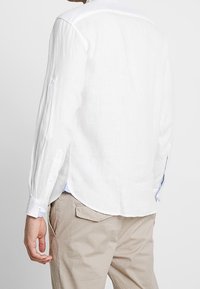 Biała lniana koszula z długimi rękawami, z guzikami przy mankietach i zaokrąglonym dołem. Posiada subtelne teksturowane wykończenie oraz jasnoniebieskie akcenty przy kołnierzu.