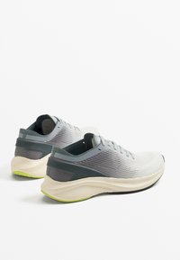 Szürke és fehér sportcipők texturált hálós felsőrésszel, párnázott szegéllyel és vastag, ívelt talppal, amely zöld díszítést tartalmaz.