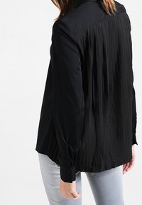 Black long-sleeve blouse with a pleated back design, made of smooth fabric. Features buttoned cuffs and a classic collar.