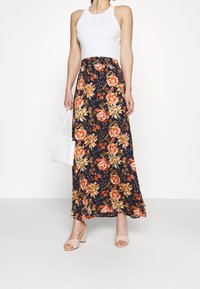 Robe maxi avec un motif floral bleu marine mettant en avant des roses orange et des touches jaunes. Accompagnée d'un sac blanc et de talons beiges.