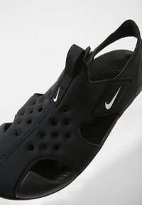 Zapatilla deportiva negra con un diseño ligero, que presenta orificios cortados para ventilación, una correa para un ajuste seguro y un acento de logo visible.