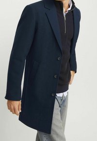 Marinblå överrock med skräddarsydd design, fem knappar och sidofickor, lager-på-lager över en mörk tröja och en ljus skjorta med krage.