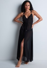 Robe maxi noire transparente avec un décolleté en V profond, des bretelles ajustables, des accents dorés, une taille à nouer et une fente haute sur la cuisse. Tissu lisse et léger.