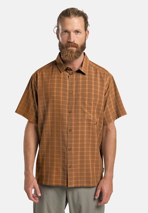Man met baard die een bruine geruite korte mouw blouse en lichte broek draagt, staand tegen een effen achtergrond.