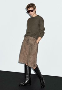 Tmavě zelený svetr, hnědé semišové kalhoty po kolena a vysoké černé kožené boty. Model má na sobě oversized sluneční brýle a stojí na světlém pozadí.