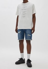 Světlé šedé grafické tričko s krátkým rukávem, doplněné modrými denimovými šortkami s vybledlými detaily a ohrnutými manžetami. Černé tenisky a bílé ponožky.