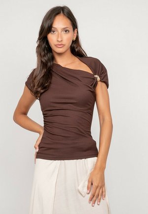 Femme aux longs cheveux foncés portant un haut marron à manches courtes avec une boucle décorative et une jupe beige clair, posant devant un fond neutre.