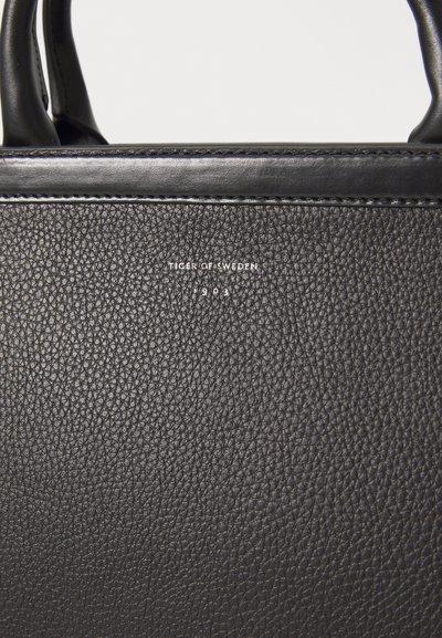 Schwarze Lederhandtasche mit strukturiertem Oberfläche, mit geprägtem Branding "TIGER OF SWEDEN" und einer glatten Kante oben.
