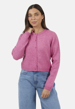 Femme aux cheveux longs bruns portant des boucles d'oreilles dorées en forme d'anneau, un cardigan rose boutonné et un jean bleu, posant avec une main près de son cou.