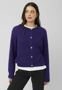 Cardigan violet tricoté avec texture côtelée, encolure ronde, boutons argentés sur le devant, et manches longues, porté sur une chemise blanche.