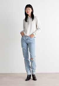 Lichtgrijze hoodie met een trekkoord, gedragen met lichtblauwe distressed jeans met kniebels. Zwarte enkellaarzen maken de outfit compleet.