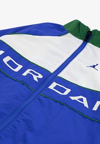 Vēja necaurlaidīga jaka ar zilu audumu, baltām un zaļām akcentiem. Aprīkota ar priekšpusi ar rāvējslēdzēju, lielu "JORDAN" tekstu un logo dizainu.