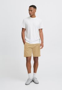 Tricou alb din bumbac, pantaloni scurți khaki și adidași gri cu șosete albe. Pantalonii scurți sunt rulați la margine; ținută casual, design minimal.