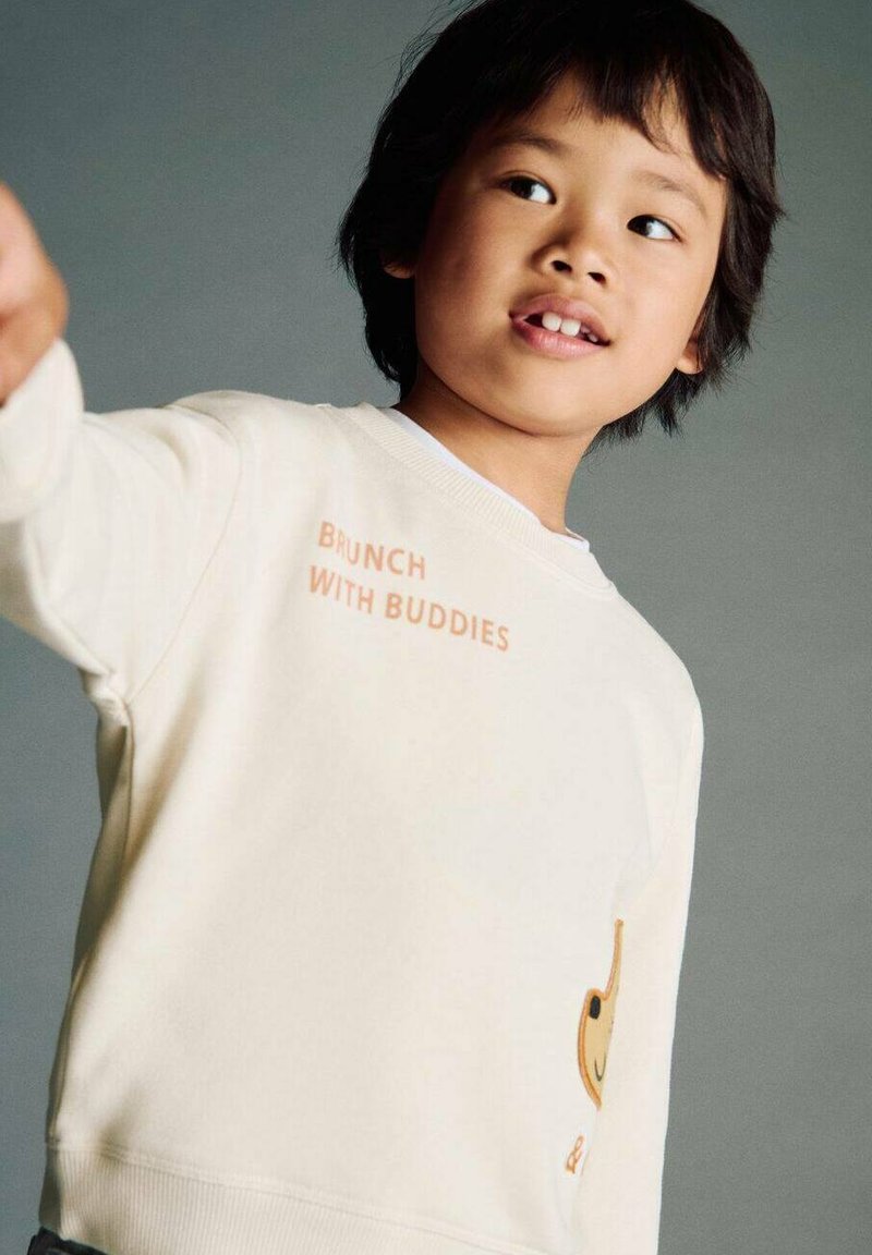 Sweatshirt color crema chiaro con la scritta "BRUNCH WITH BUDDIES" stampata in arancione, caratterizzato da un design giocoso di un personaggio sorridente di lato.
