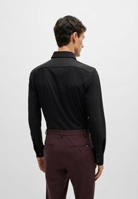 Schwarz langärmliges Hemd mit klassischem Kragen, das eine glatte Textur und geknöpfte Manschetten aufweist, kombiniert mit kastanienbraunen Hosen, die über Gesäßtaschen verfügen.