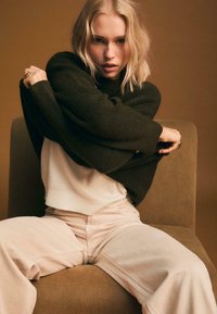 Cremefarbener Pullover unter einem dunkelgrünen, übergroßen Strickpullover, dazu weite, blassrosa Hosen. Das Model sitzt auf einem braunen Stuhl vor einem gedämpften Hintergrund.