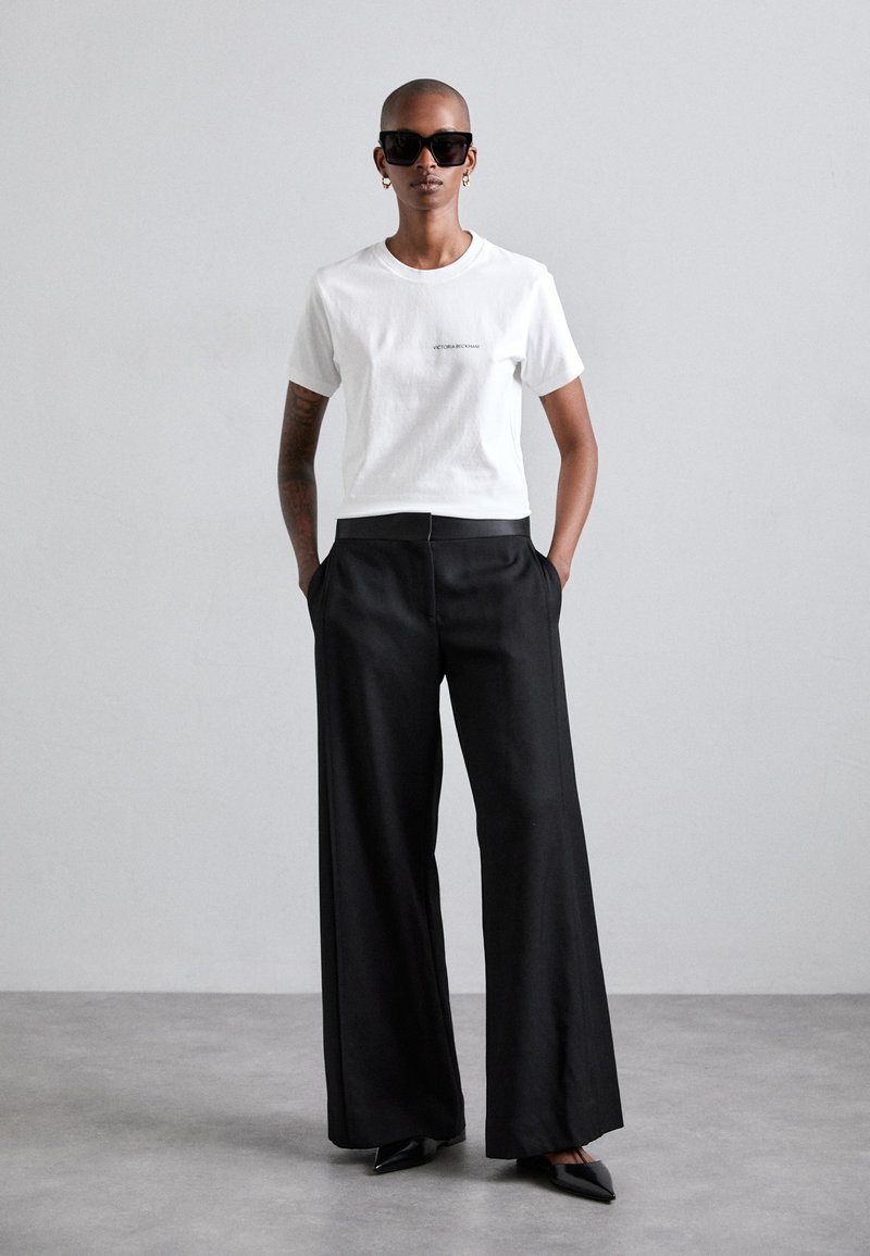 Camiseta blanca de manga corta con texto de la marca, combinada con pantalones largos de pierna ancha negros y tacones de punta negra; accessorios con gafas de sol.