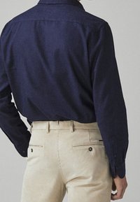 Man met een donkerblauw overhemd met lange mouwen, ingestoken in beige broeken, gezien vanachter tegen een effen achtergrond.