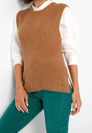 Femme portant un pull sans manches camel en maille sur une chemise blanche boutonnée et un pantalon bleu sarcelle, debout devant un fond blanc.