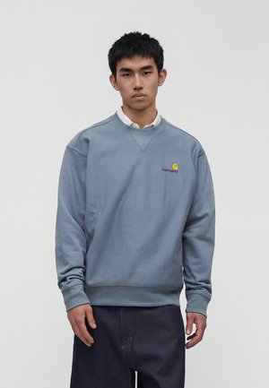 Ung mand med kort sort hår iført en blå Carhartt sweatshirt over en hvid skjorte med krave mod en ensartet lys baggrund.