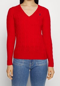 Pull rouge en maille torsadée avec un col en V, des poignets et un ourlet côtelés, arborant un logo bleu sur le côté gauche de la poitrine. Associé à un jean bleu ajusté.