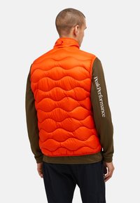 Gilet imbottito arancione con un design trapuntato. Presenta un colletto alto e una camicia a maniche lunghe marrone a contrasto con la scritta "Peak Performance" stampata.