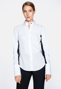 Camicia bianca con bottoni, caratterizzata da pannelli laterali scuri a contrasto, maniche lunghe con polsini allungati e un classico design del colletto.