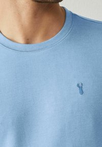 Maglietta di cotone blu chiaro con scollo rotondo e un piccolo motivo ricamato a forma di aragosta in filo abbinato sul lato sinistro del petto.