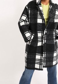 Cappotto a quadri bianco e nero in misto lana con maniche lunghe, tasche frontali e colletto a rever. Abbinato a jeans in denim blu.