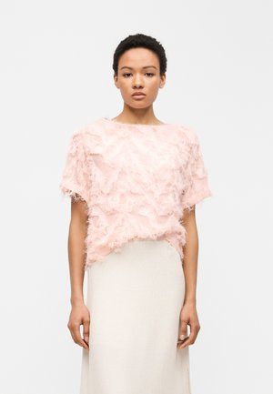 Γυναίκα με κοντά κατσαρά μαλλιά που φοράει μπλούζα με κοντά μανίκια σε απαλό ροζ με υφή και φούστα υψηλής μέσης σε εκρού, μπροστά σε μονόχρωμο φόντο.