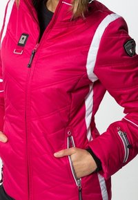 Różowa pikowana kurtka z białymi akcentami, kieszeniami na zamek i łatką z logo. Posiada wysoki kołnierz oraz trwały, gładki materiał.