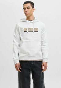 Witte hoodie met trekkoorden, voorzak en een rechthoekig logo in beige en zwart. Gecombineerd met donkere spijkerbroek. Eenvoudig ontwerp en zachte stof.
