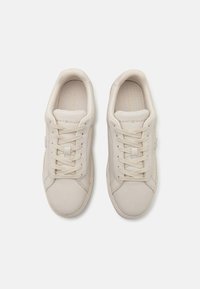Light beige sneakers with smooth leather upper, flat laces, stitched detailing, and logo on the tongue. Cushioned interior and rounded toe.